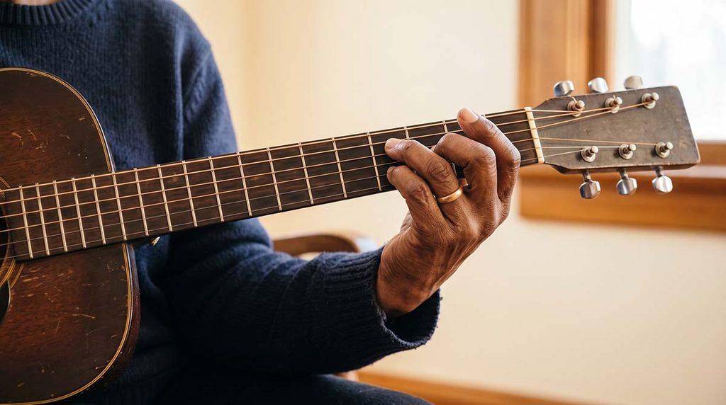 Older woman’s fingertips pressing guitar strings, showing the physical difficulty that meaningful songs don’t remove